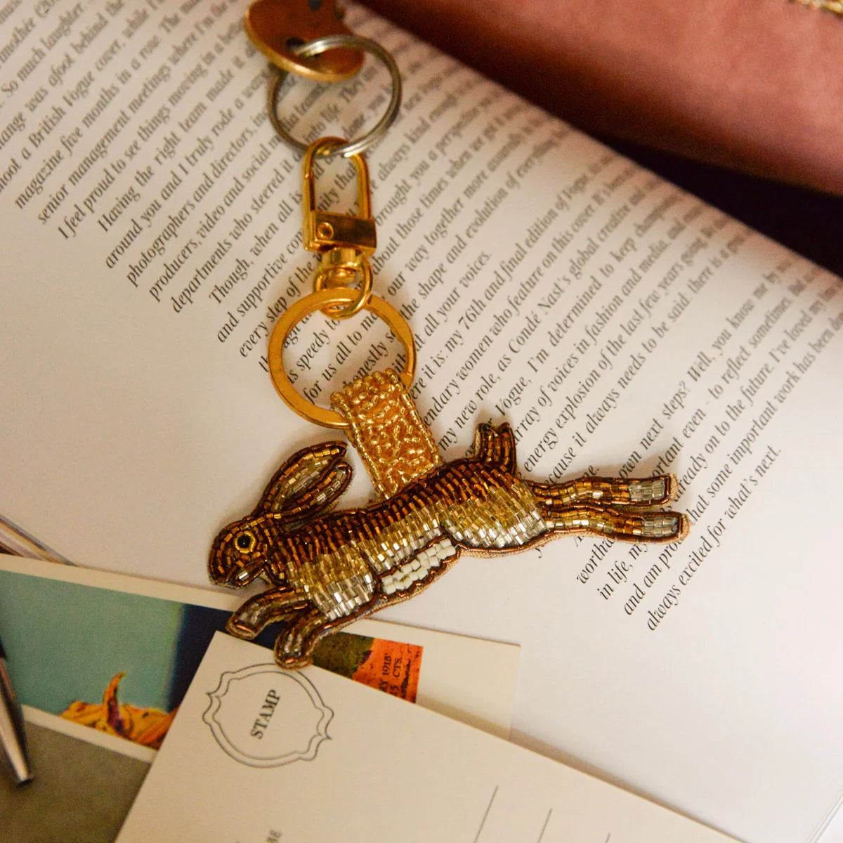 A gold and brown sequinned rabbit keyring rests on an open book, with a postcard and a pink bag partially visible nearby.