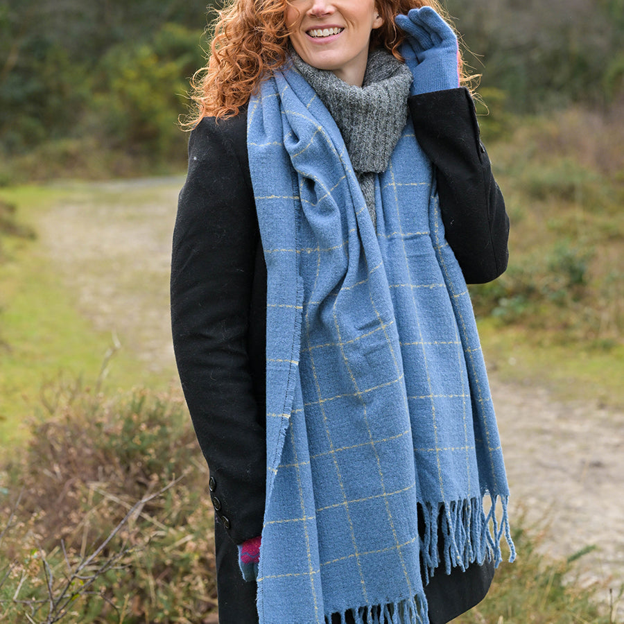 Woman wearing a light purple broderie anglaise dress and a colourful patterned scarf sits outdoors, smiling, with greenery in the background.