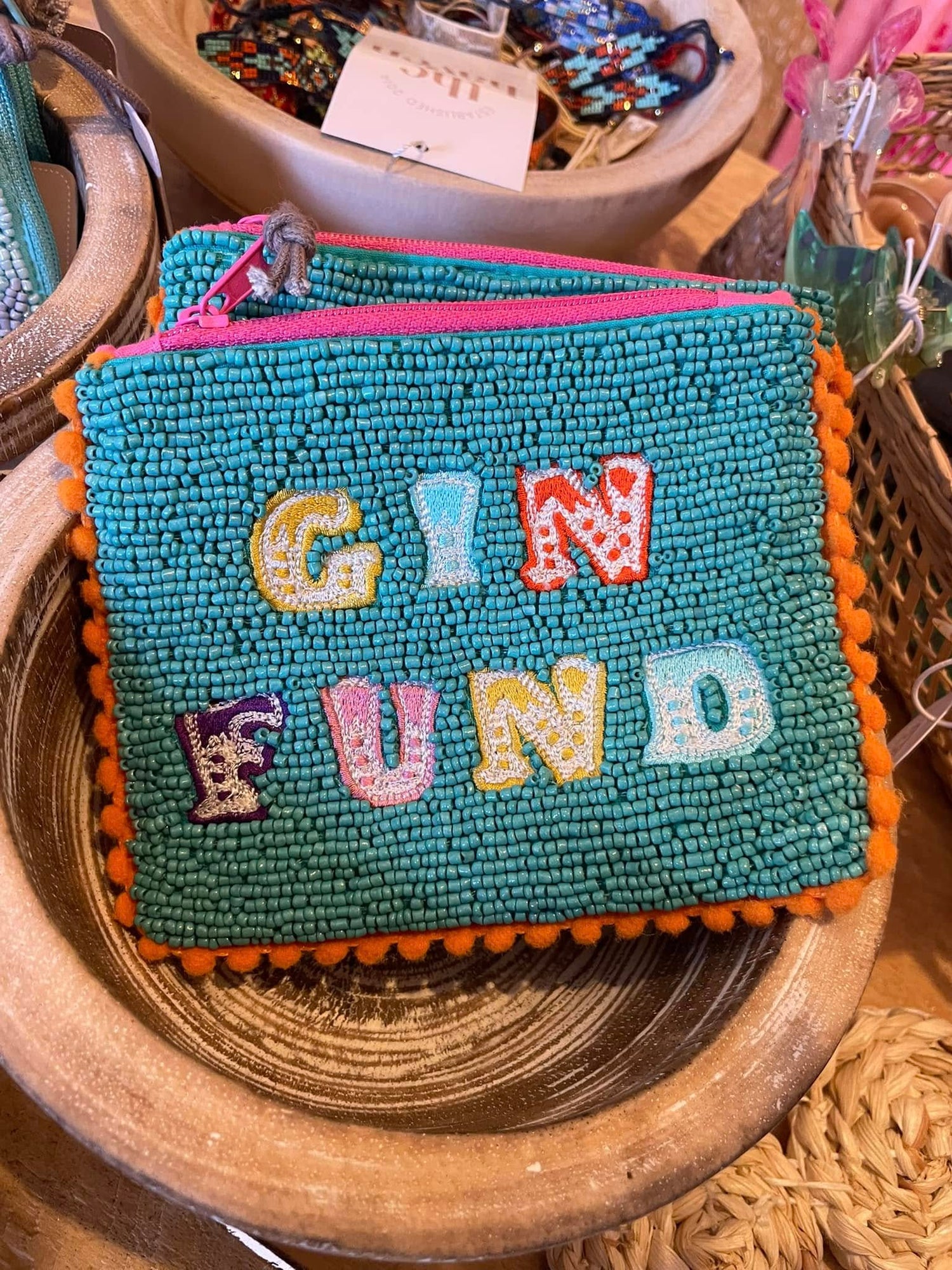 A beaded pouch with the words GIN FUND in colourful letters sits in a shallow wooden bowl among other decorative items.
