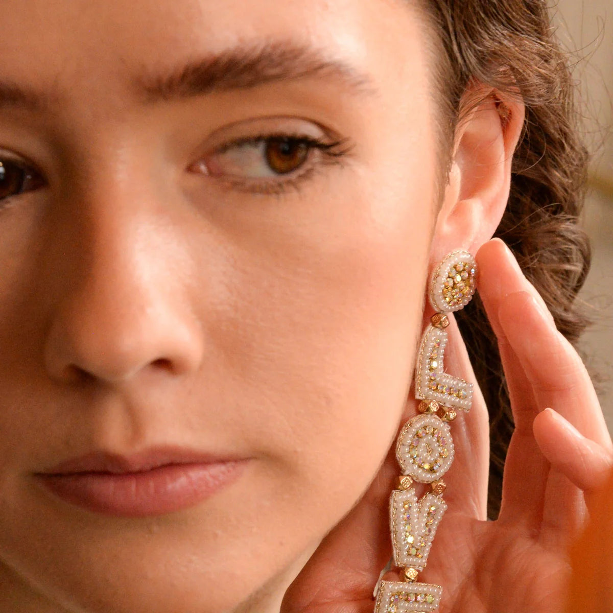 A person with brown eyes and wavy hair wears a large gold and pearl earring spelling out the word LOVE while touching it with their hand.