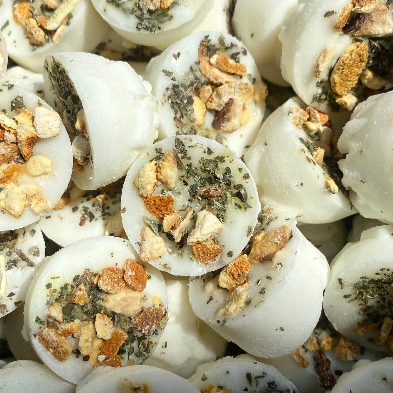 Round, white bath melts topped with dried herbs and citrus peel, closely arranged in a pile and illuminated by natural light.