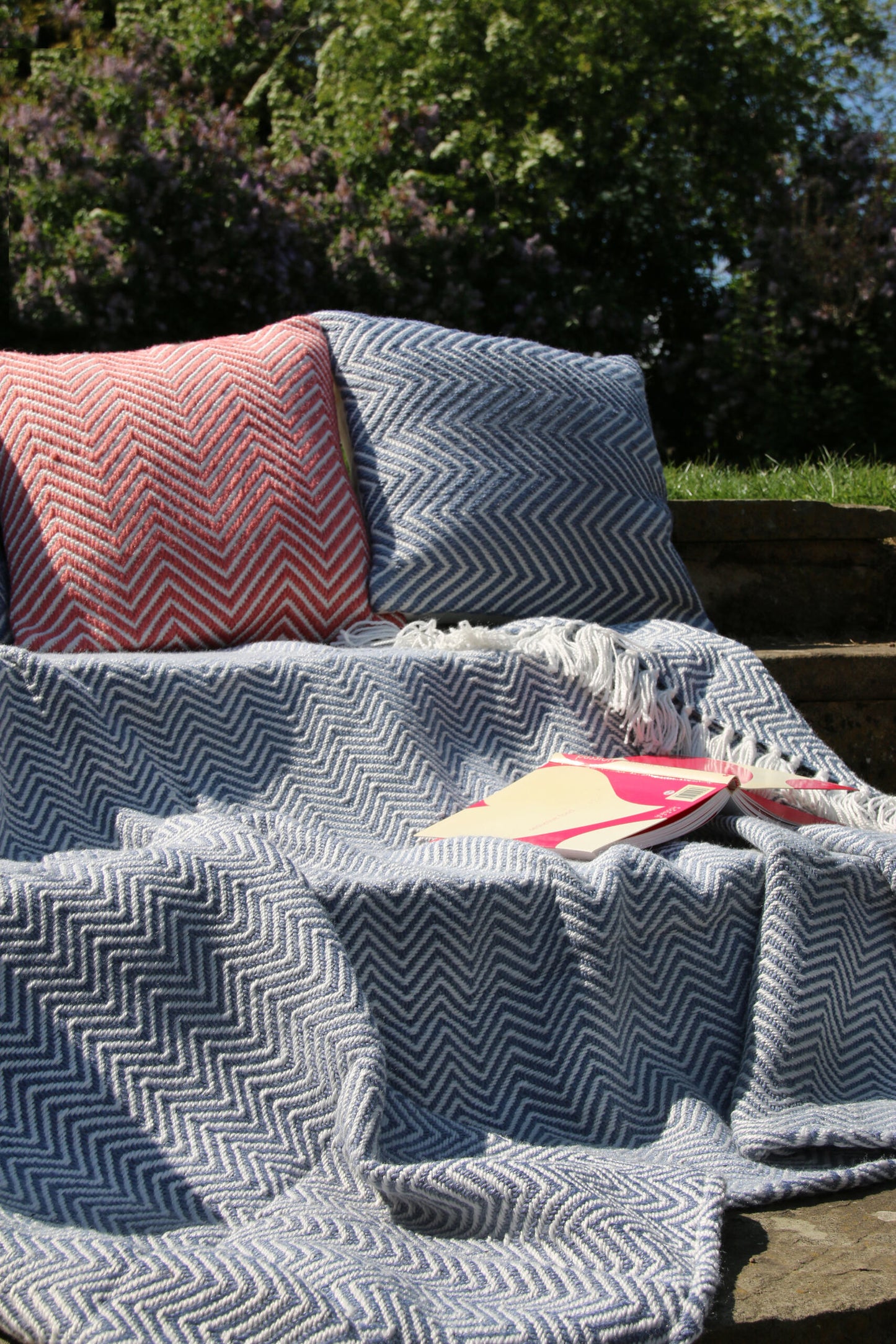 Two cushions and a blanket with a zigzag pattern are arranged on outdoor steps, alongside a notebook featuring a pink and yellow cover. Sustainable rugs crafted from recycled plastic bottles enhance the scenes charm, whilst greenery flourishes in the background.