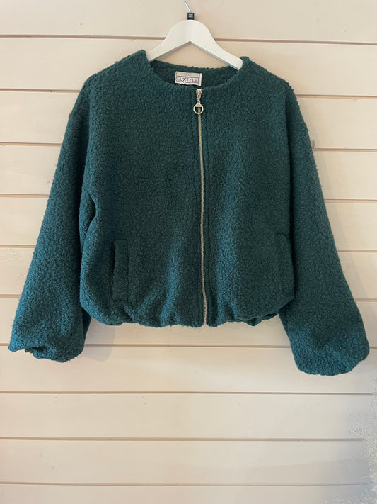 A teal, zip-up, long-sleeved, sherpa vegan bomber jacket hangs on a white hanger against a white wooden wall, ideal for those who appreciate sustainable fashion.