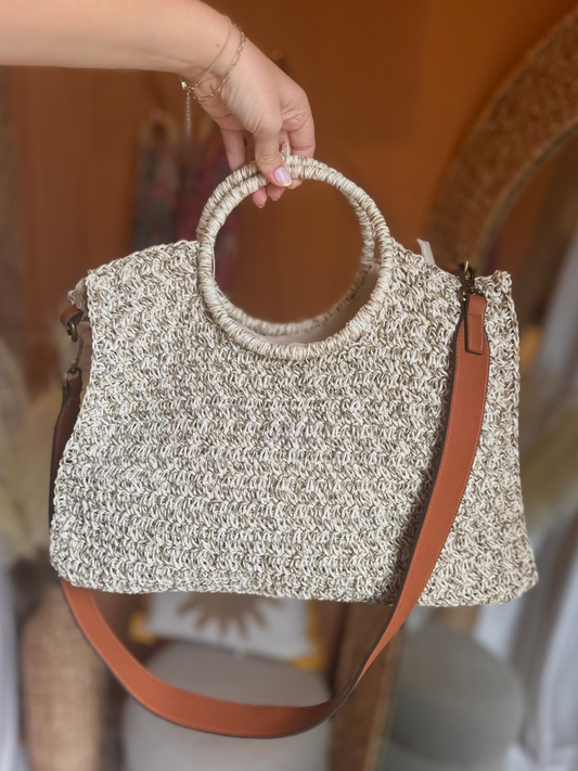 A hand holds a woven beige handbag with circular handles and a brown shoulder strap against a blurred indoor background.