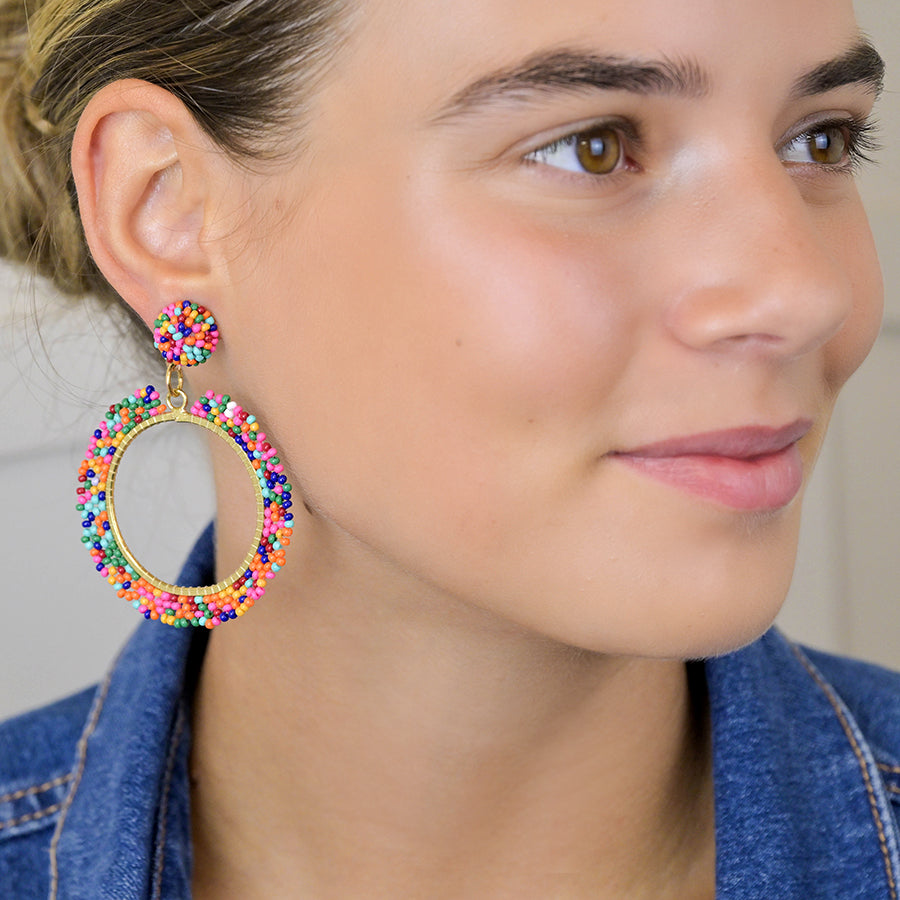 Individual wearing vibrant mixed bead earrings with faux gold plating and a denim shirt, smiling slightly while looking to the side.