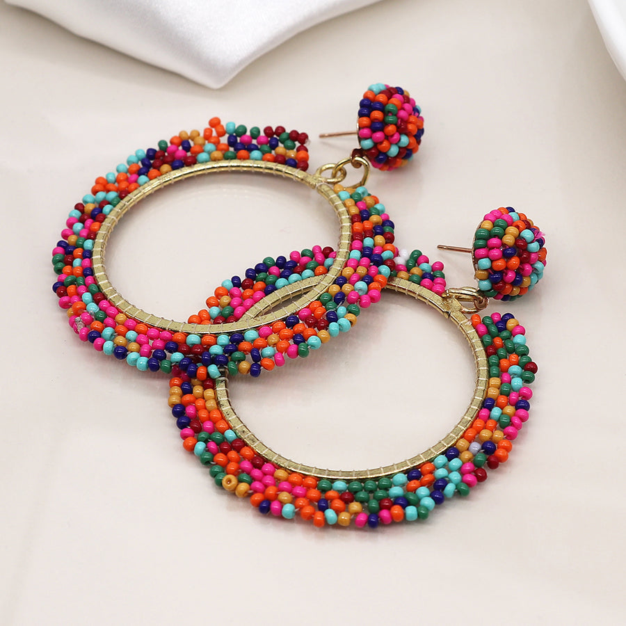 Vibrant mixed bead earrings with colourful beaded hoops and round studs, featuring faux gold plating accents, rest beautifully on a light surface.