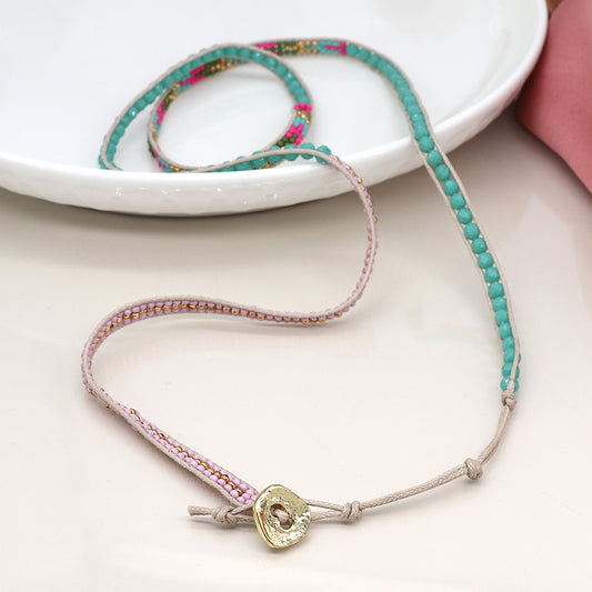 A beaded necklace, which also serves as a wraparound bracelet with turquoise, pink, and gold beads, tied with beige thread and finished with a round, golden button fastening, is placed on a white surface.