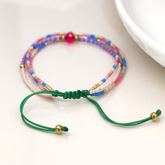 The Pink & Blue Mix Adjustable Beaded Bracelet, a sophisticated accessory adorned with pink, blue, and gold beads, is secured with luxurious green strings and rests elegantly on a white surface.