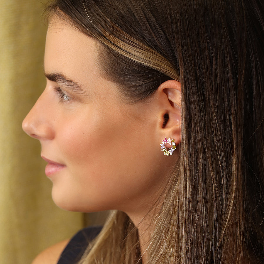 A woman with long brown hair is wearing pink and gold floral circle studs, facing left against a beige background.