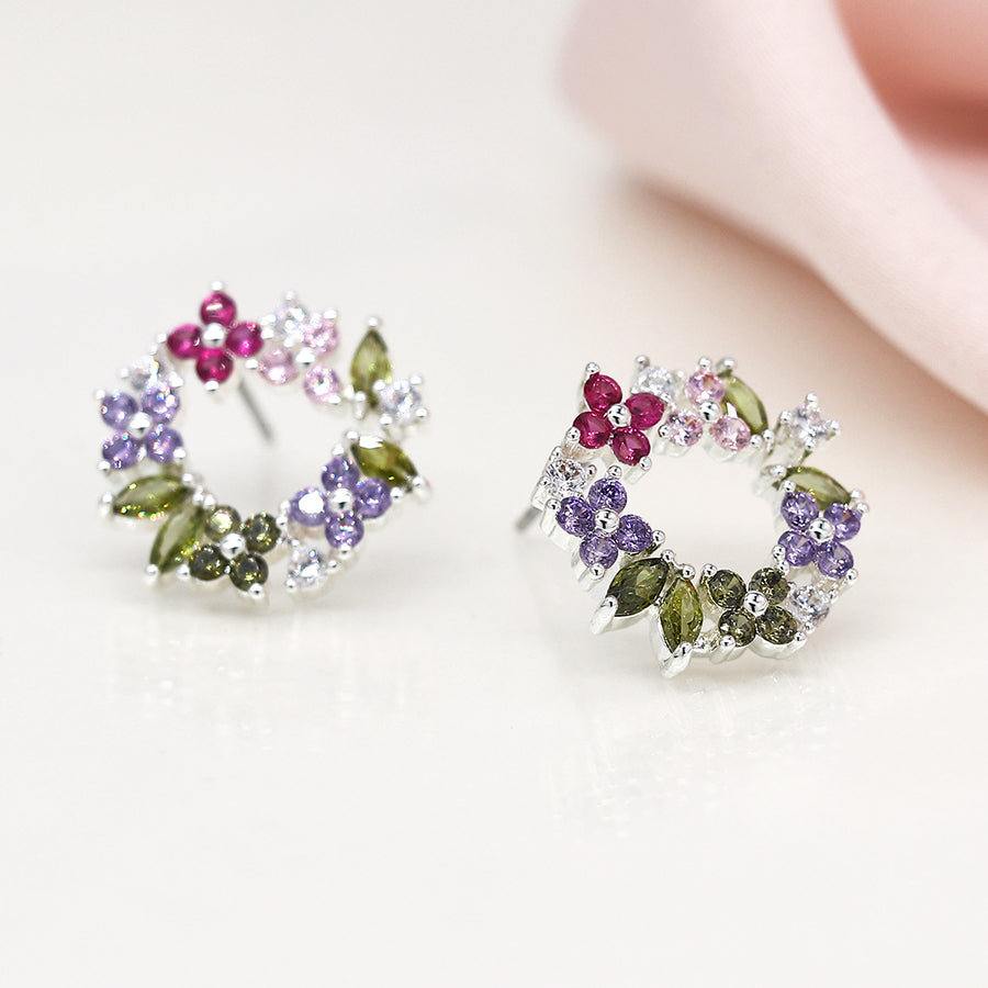 Two floral circle stud earrings, silver-plated and adorned with faceted crystals in pink, purple, and green hues, shimmer against a soft, out-of-focus background.
