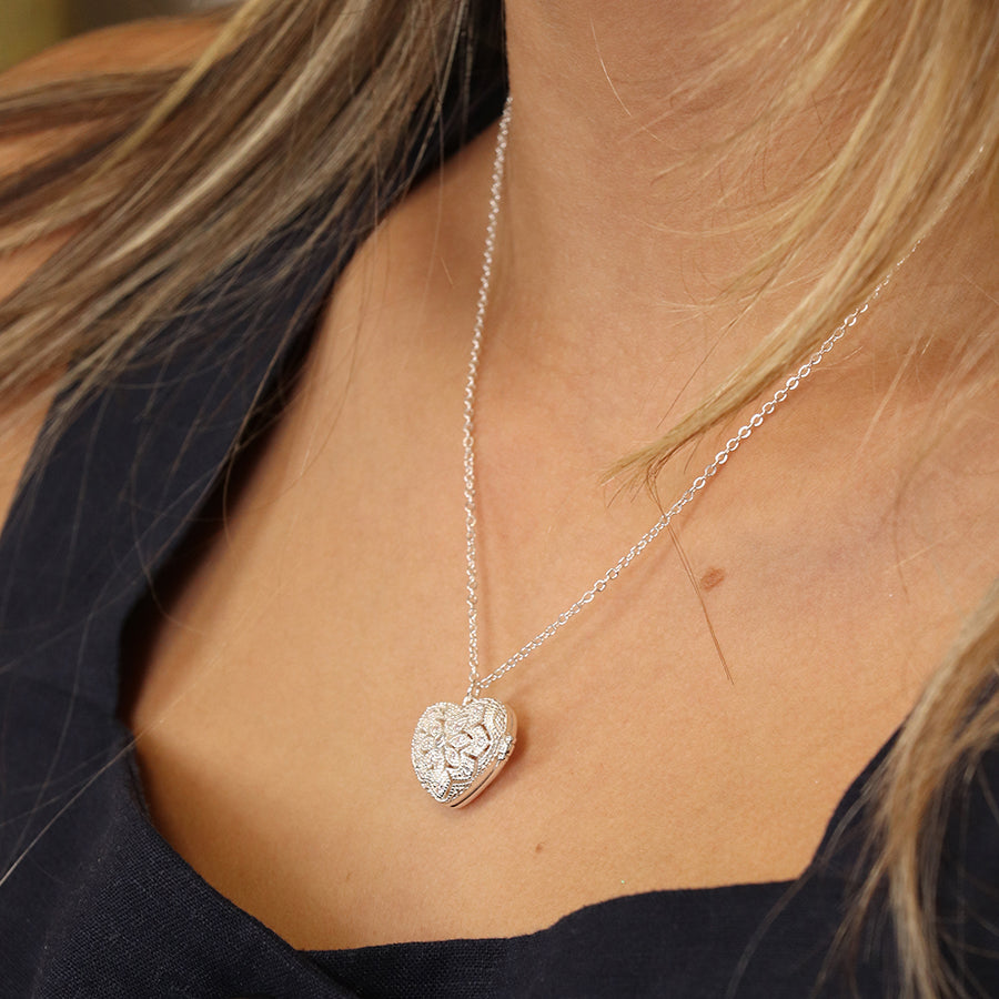 A person wearing a silver-plated, heart-shaped pendant with intricate detailing on a chain necklace, displayed as part of their exquisite jewellery collection against a dark top.
