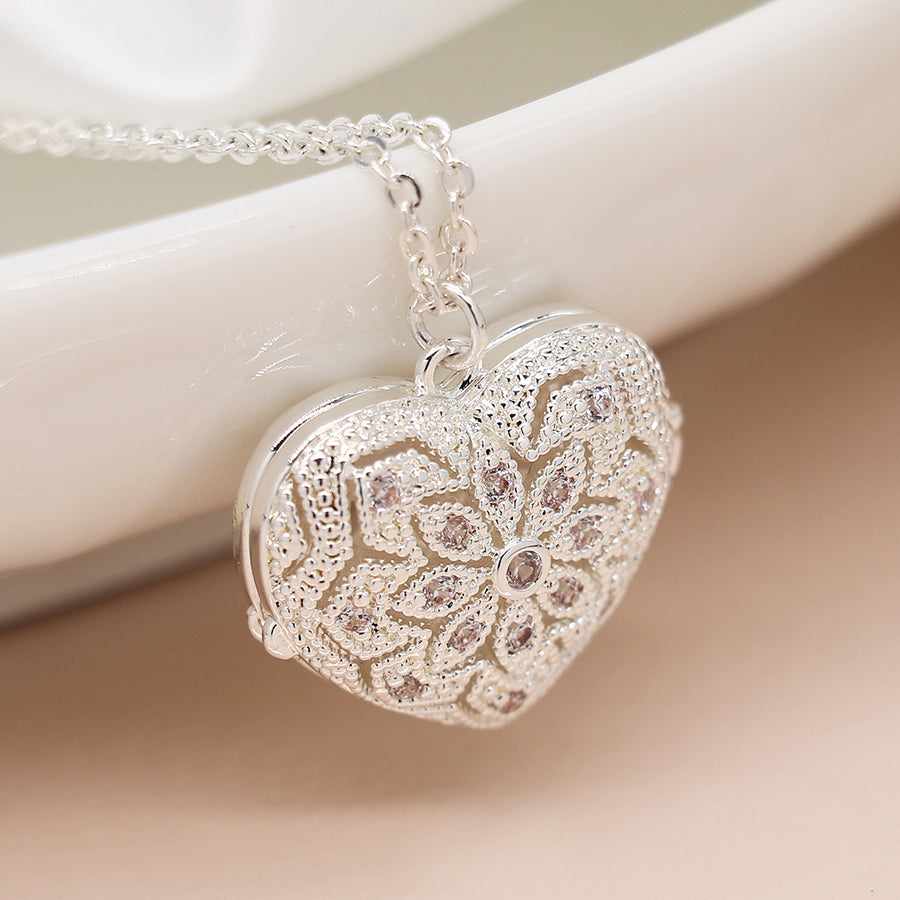 A silver-plated heart-shaped locket with an intricate floral pattern hangs from a chain, resting on a white ceramic surface, adding elegance to any jewellery collection.