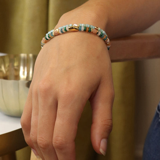 A close-up of a persons hand displaying a luxury accessory—a beaded bracelet crafted with teal and light pink beads—resting elegantly on a wooden surface.
