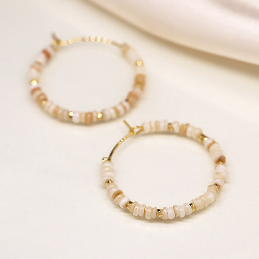 Close-up of two faux gold-plated hoop earrings adorned with small beige and white beads arranged in a circular pattern, resembling seed pearls, placed on a light-coloured surface.