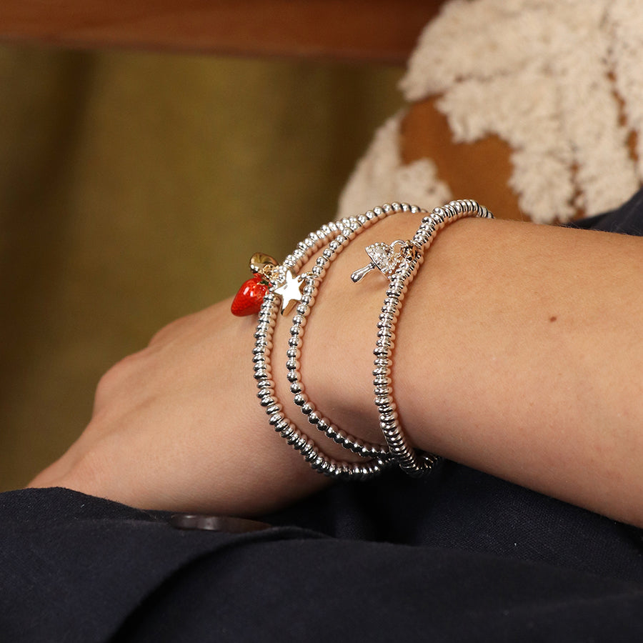 A wrist adorned with three silver-plated charm bracelets exudes edgy elegance, featuring tiny treasures such as a strawberry, a bow, and a whimsical toadstool charm.
