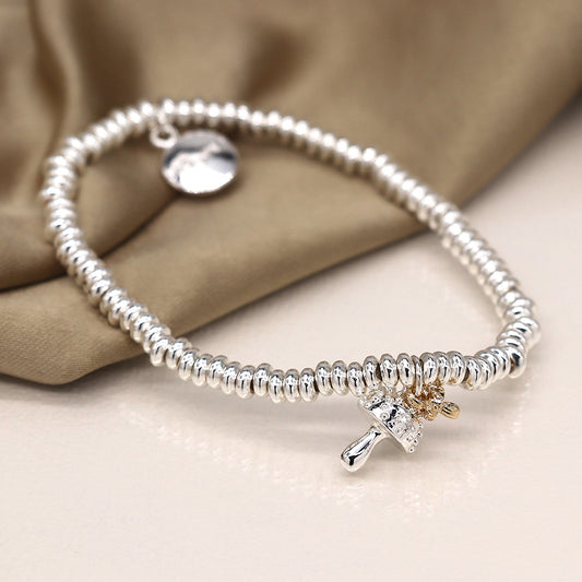 A silver-plated charm bracelet with a toadstool charm and a small round tag rests on a soft beige fabric background, exuding an air of edgy elegance.
