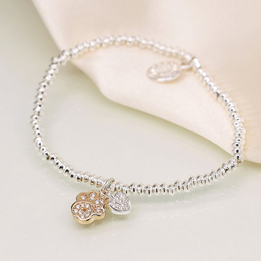 A silver-plated charm bracelet adorned with a golden paw charm and a crystal heart, beautifully resting on a light-coloured surface.
