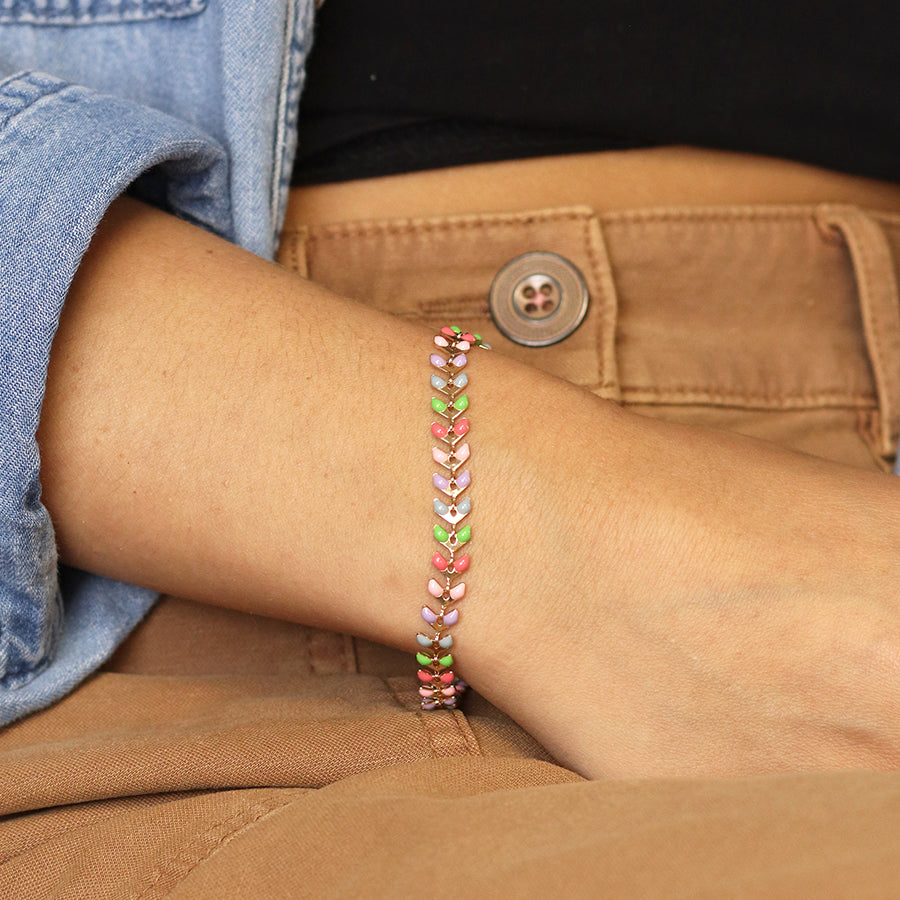 A close-up of a person wearing the Golden Pastel Epoxy Droplets Bracelet, a luxurious accessory featuring leaf-shaped links. They are dressed in a light blue denim shirt and brown trousers with a visible button.