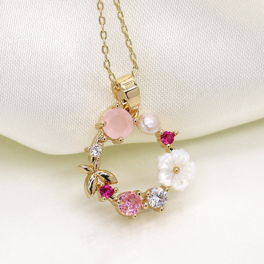 Elegant faux gold-plated necklace with a circular pendant featuring pink and white gemstones, a pearl, and a small gold leaf on a soft white background, reminiscent of delicate shell flowers.