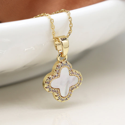 A statement piece, this gold necklace with a clover-shaped pendant features a white centre surrounded by small clear stones, resting elegantly on a white ceramic surface.