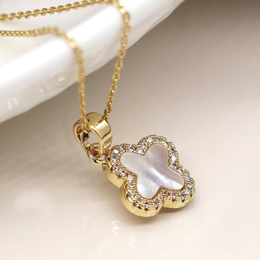A statement piece, this necklace features a faux gold chain and a pendant shaped like a four-leaf clover, boasting a mother of pearl centre surrounded by small clear stones on a white surface.