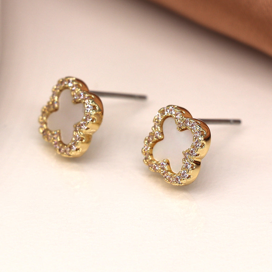 Imitation gold-plated clover-shaped studs, adorned with small gemstones, shimmer elegantly on a smooth, light-coloured surface.
