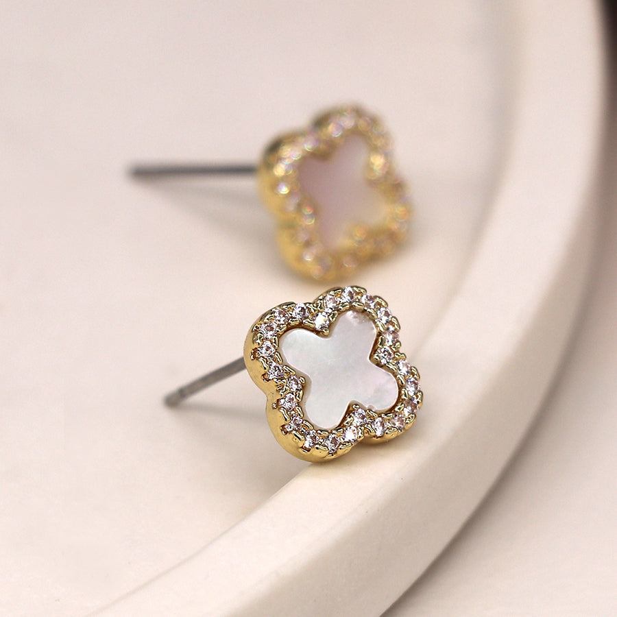 Two faux gold-plated earrings with a floral design, featuring white and light pink centres, surrounded by small clear stones, placed on a soft white surface.