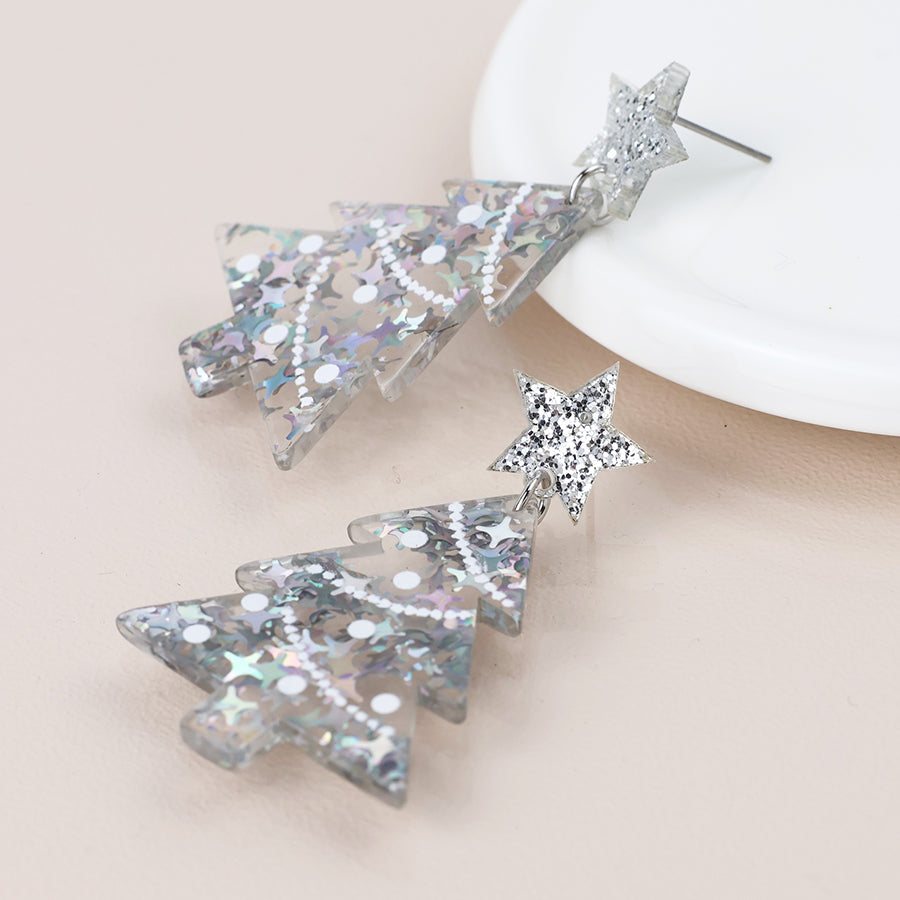 Two silver glitter acrylic Christmas tree earrings with star-shaped studs, displayed on a light surface beside a white dish—a festive touch of holiday jewellery.
