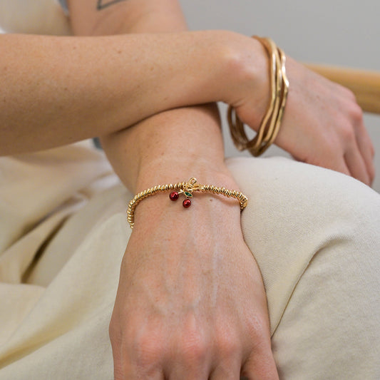A person wearing a gold bracelet adorned with cherry charms and several gold bangles rests their arm across their lap.