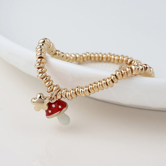 A gold beaded ring with a small red toadstool charm and star sits on a white ceramic dish.
