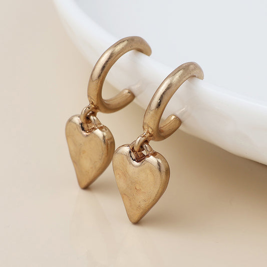 A pair of small gold earrings with dangling heart-shaped charms, evoking a vintage style, displayed beside a white dish.