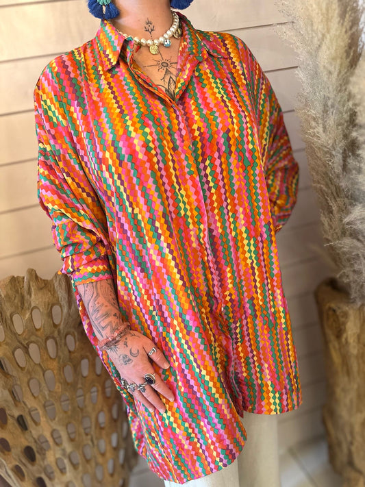 Person wearing a Loxy Long Top—a loose, long-sleeved shirt featuring a multicoloured geometric pattern and made from vegan-friendly materials—showing tattoos and rings while posing indoors near dried plants.