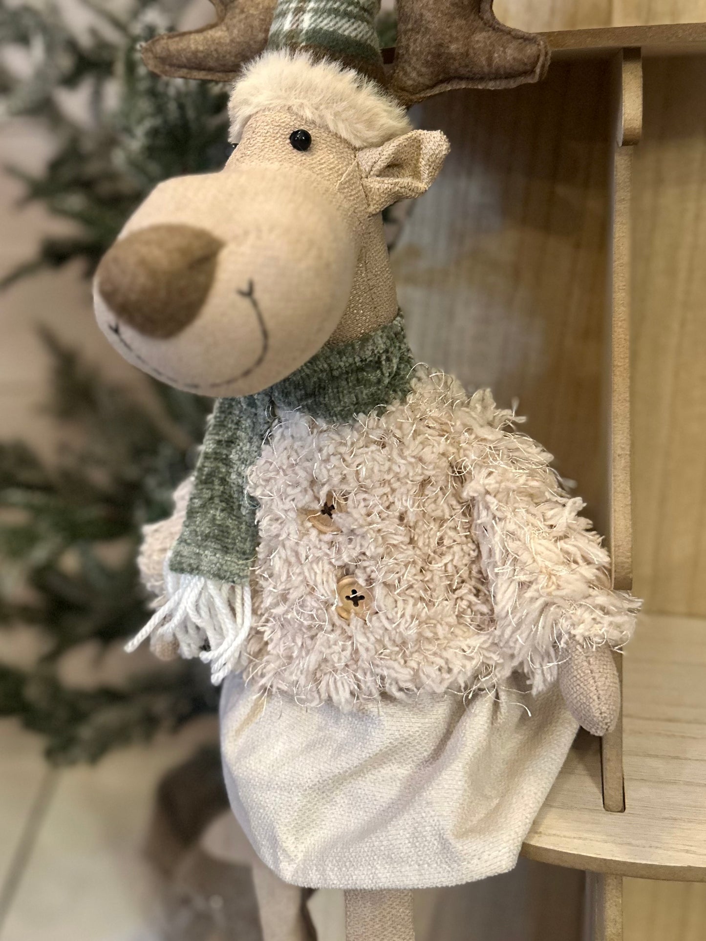 A plush Dangly Legs Christmas Deer Decoration dressed in a green scarf, fluffy cream jumper, and white skirt, seated on a wooden surface—ideal as a charming festive décor accent.