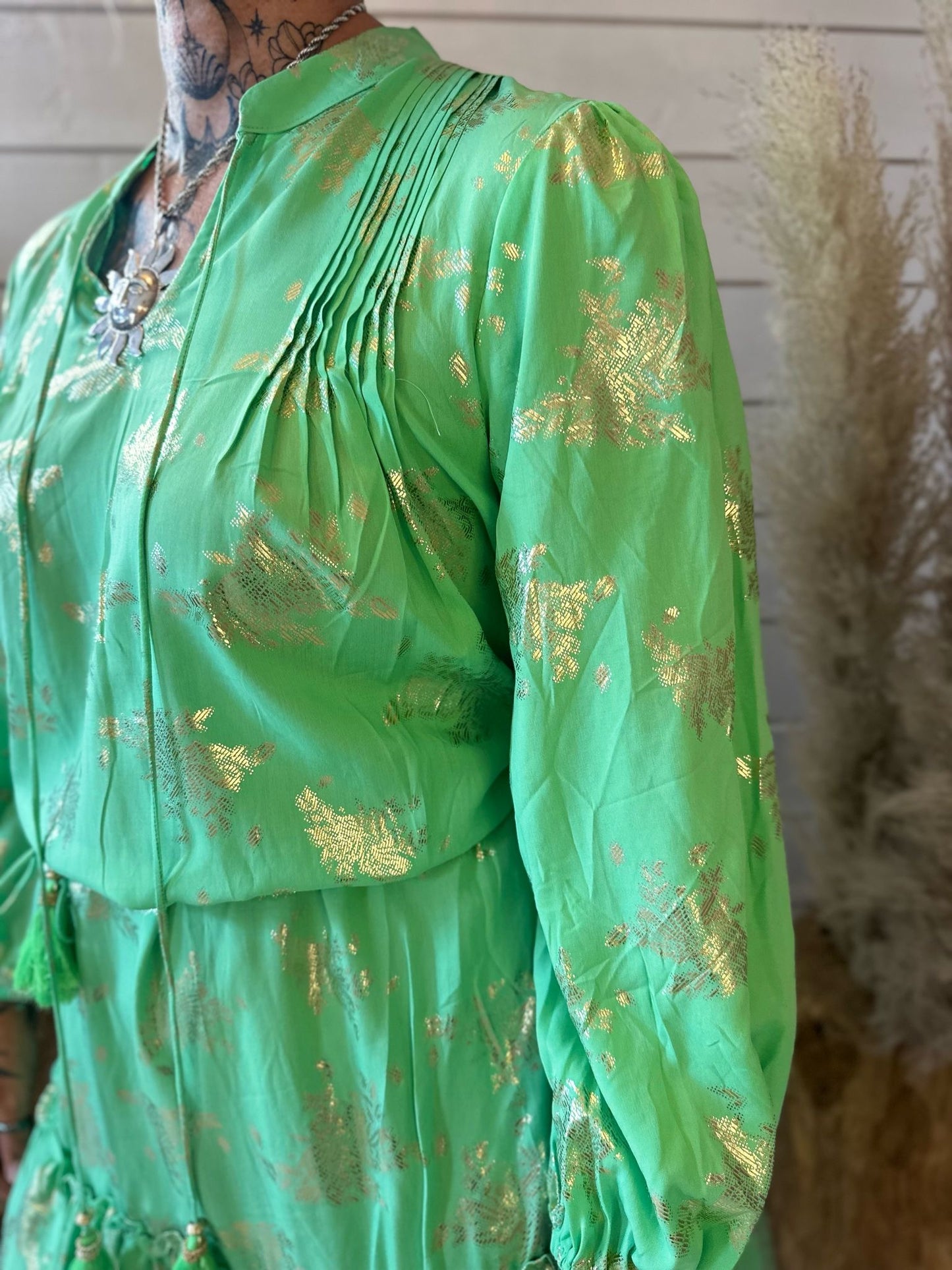 Person wearing the chic and stylish Green Gold Smock Dress with gold patterns and chest pleats, featuring a necklace and visible neck tattoos. Neutral background with dried plants. *British English Version:* Individual wearing a chic and stylish green-and-gold smock dress adorned with gold patterns and pleating at the chest, complemented by a necklace and visible neck tattoos. Neutral backdrop with dried foliage.