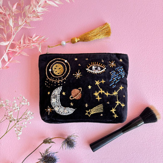 A black velvet make-up pouch adorned with celestial bead embroidery and a gold tassel rests on a pink surface, surrounded by a make-up brush and decorative dried flowers.