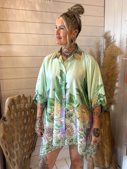 Woman with tattoos stands indoors wearing a loose, light green patterned shirt featuring botanical and animal designs, looking to the side.