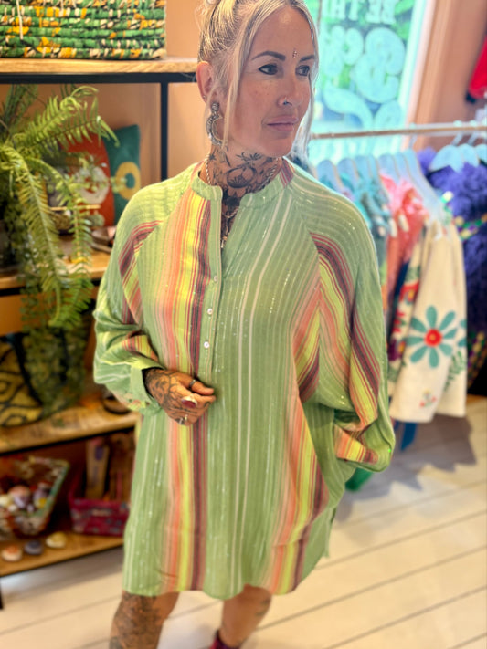 A woman with tattoos wears a loose, striped green dress—an effortlessly stylish piece—and black boots, standing indoors near shelves with plants and colourful decor.