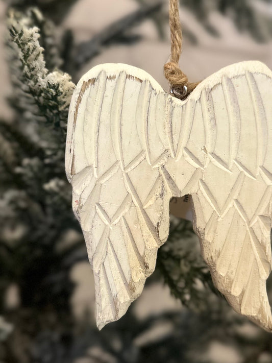 A white wooden angel wing decoration hangs by a string before a snow-dusted pine branch, bringing a festive touch to your Christmas tree.