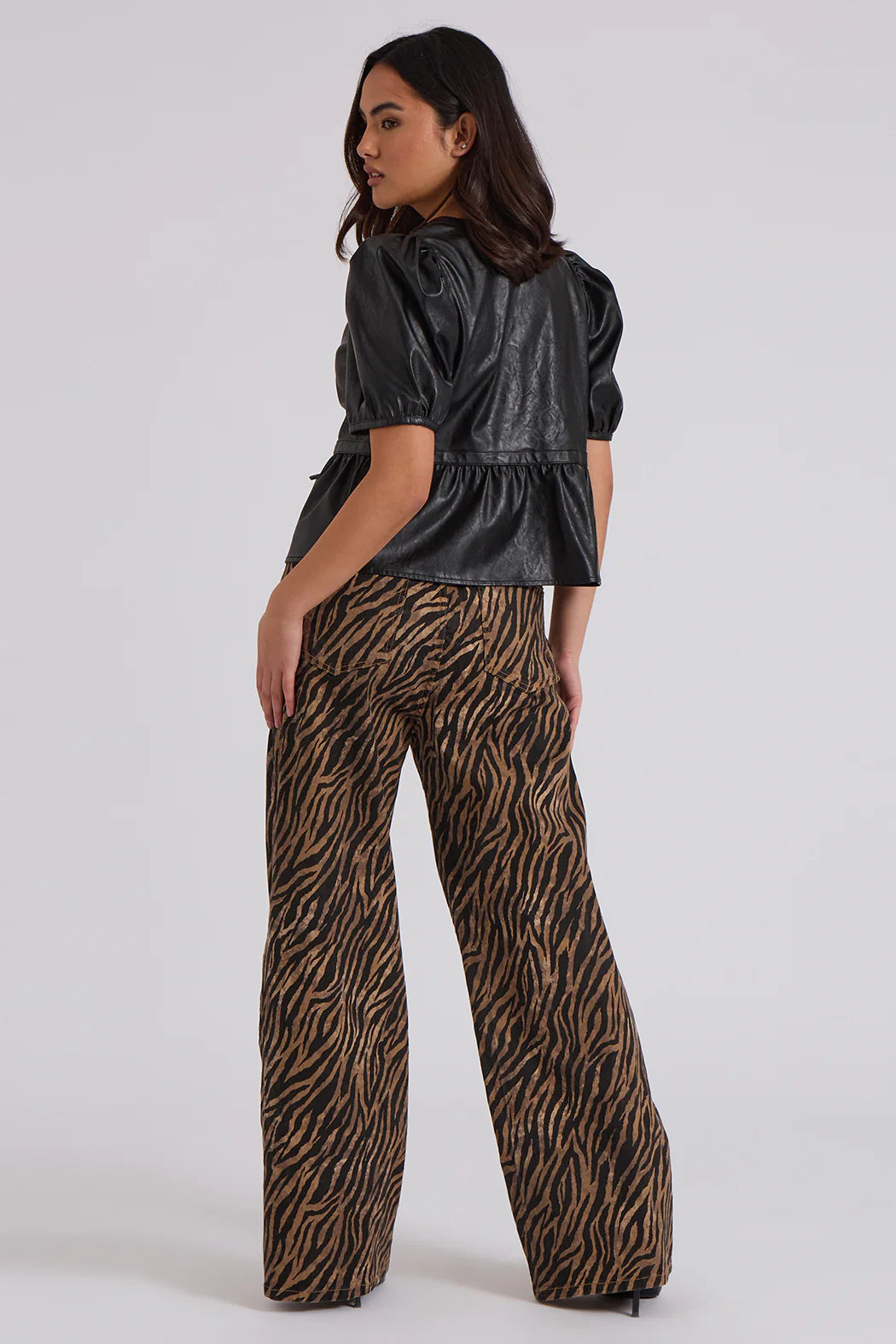 A person stands with their back turned, wearing a short-sleeved black leather top and high-rise, wide-leg trousers featuring a bold zebra print pattern over a light grey background.
