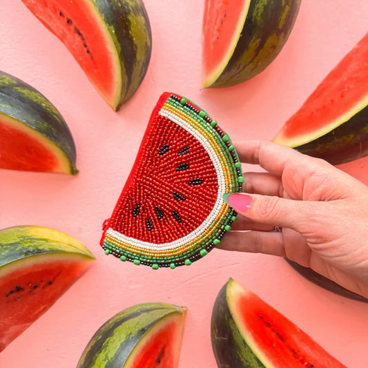A hand holds a Watermelon Purse, a vegan-friendly accessory shaped like a watermelon slice, surrounded by fresh watermelon wedges on a pink background—perfect for summery style.