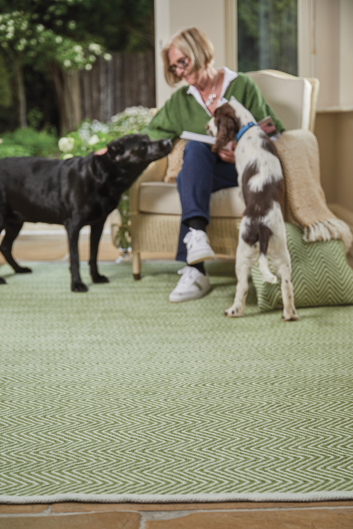 A person sits in a chair stroking two dogs, one black and the other white with brown spots, on a green patterned Herringbone Washable Rug made from recycled plastic bottles indoors.