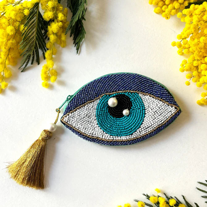 The Glimmer Eye Purse is a beaded, vegan-friendly coin purse in the shape of an eye, embellished with pearls and a gold tassel, displayed on a white surface surrounded by yellow flowers.