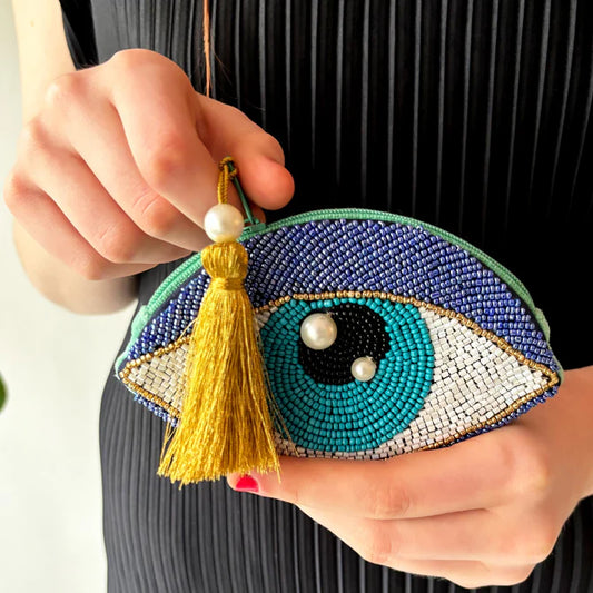 Person holding a vegan-friendly Glimmer Eye Purse—a small beaded coin purse shaped like an eye, embellished with pearls and a gold tassel, set against a backdrop of black pleated fabric.