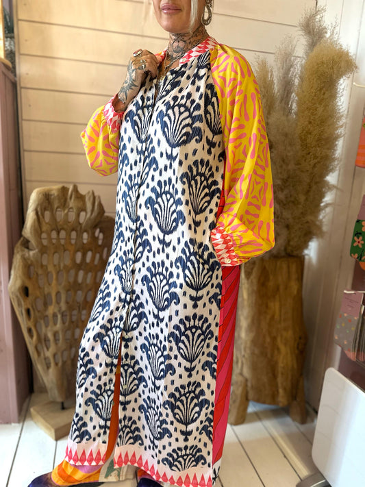 A person stands indoors wearing a long patterned dress with black and white designs, yellow and pink sleeves, and orange details, beside decorative wooden and plant elements.