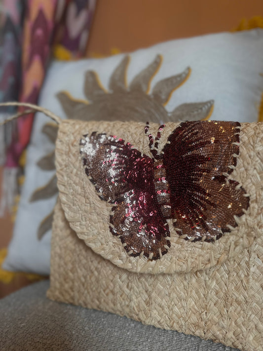 A woven straw clutch, the Red Butterfly Bag, featuring a large sequinned butterfly design, rests on a cushion adorned with a sun motif for a sophisticated look.