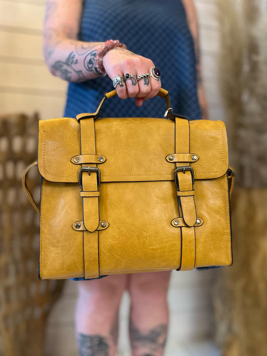 A person with tattoos holds a vegan-friendly yellow faux leather satchel by the top handle, featuring two front buckle straps that elevate their business look.