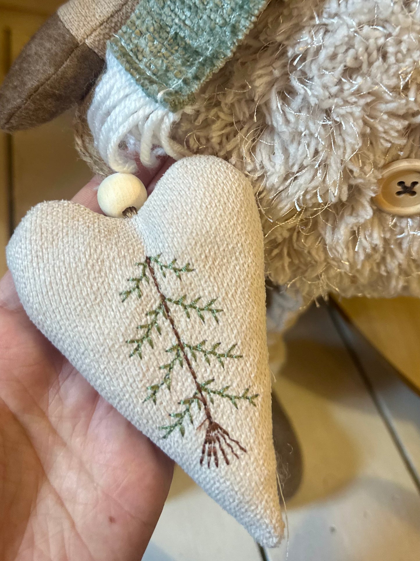 A hand holds a fabric heart ornament featuring a green embroidered branch, beside a textured soft toy with a button detail—ideal festive décor for creating a cosy atmosphere indoors or outdoors at Christmas.