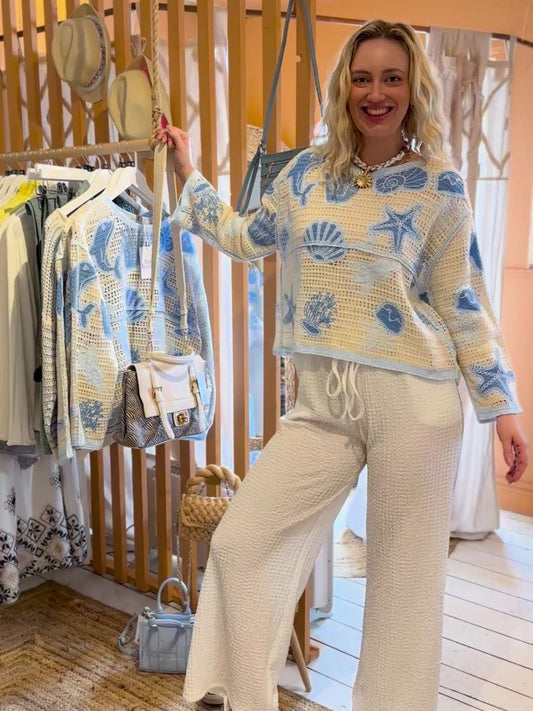 A woman stands indoors beside clothing rails, dressed in a light blue and white ocean-inspired jumper and white trousers, smiling with one hand on her hip.