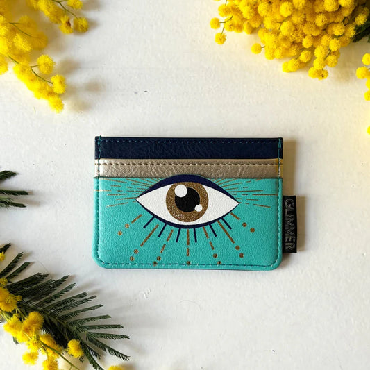 A turquoise cardholder featuring a prominent eye graphic at its centre, with gold and navy details and yellow flowers—designed as a stylish cruelty-free accessory—set against a white background.