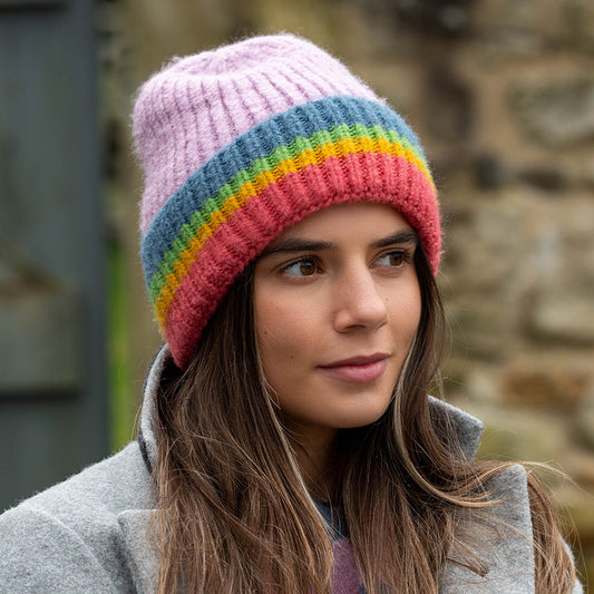 A woman wearing a grey coat and a ribbed knit beanie with rainbow stripes looks to the side outdoors.
